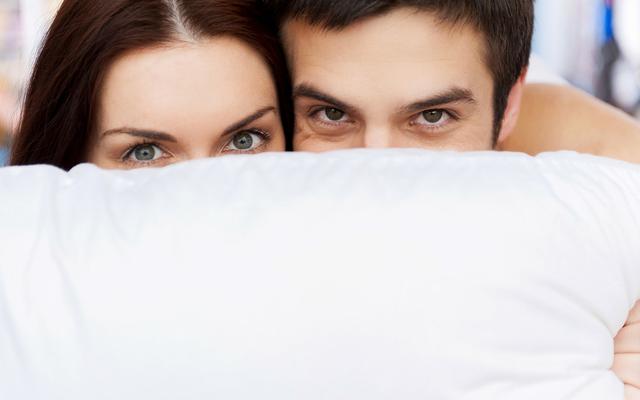 Paar im Bett versteckt sich hinter Kissen (Symbolbild)